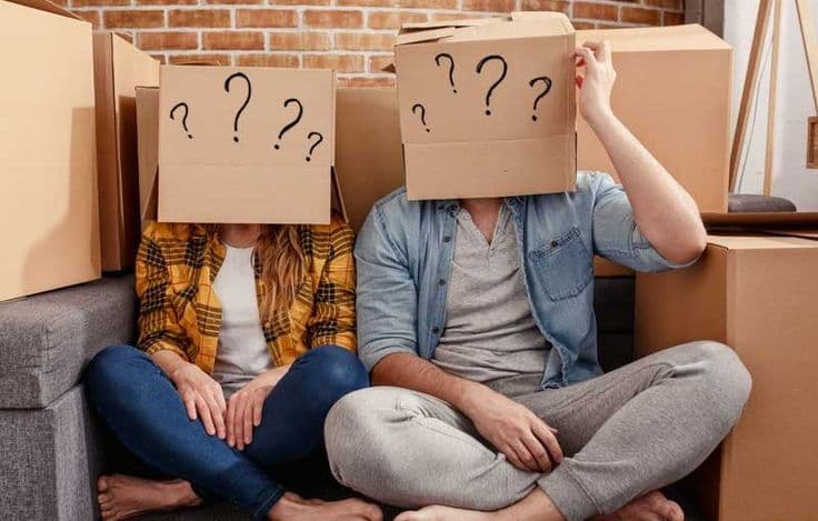Pareja sentada entre cajas de mudanza con signos de interrogación sobre la cabeza